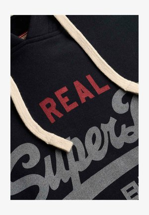 Czarna bluza z kapturem z białymi sznureczkami i czerwono-szarym napisem "REAL Superdry" nadrukowanym na tkaninie.