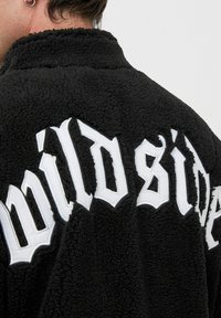 Czarna polarowa kurtka z teksturowanej tkaniny, z białym haftowanym napisem "dzika strona" na plecach.