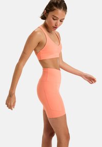 Kvinna i korallfärgad sport-bh och högmidjade cykelshorts som står på sidan, med armarna något utsträckta, mot en enkel vit bakgrund.