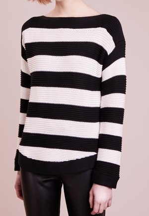 Osoba nosící černo-bílý široce pruhovaný pletený svetr a černé kožené kalhoty, zobrazena od krku po boky na světle zbarveném pozadí.