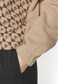 Krémová mikina s hnědými trojúhelníkovými vzory, s žebrovanými manžetami a vyšívaným detailem "ALL SZN" na rukávu. Černá látka dole.