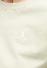 Κοντινό πλάνο ενός κρεμ πουκάμισου με μικρό λευκό κεντητό λογότυπο Jumpman στο στήθος.