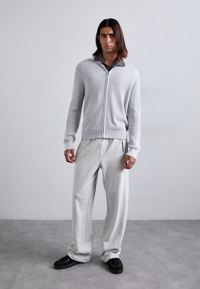 Ljusgrå ruggad zip-up kardigan över en mörk skjorta med krage, matchad med ljusgrå vida sweatpants och svarta sneakers.