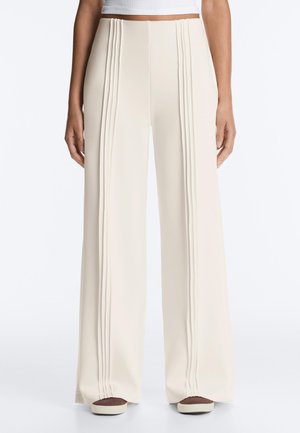 Personne portant un pantalon large crème taille haute avec des plis verticaux sur le devant, associé à des chaussures marron et blanches.