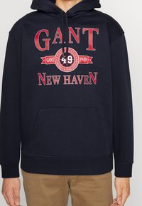 Sudadera de color azul marino hecha de una mezcla de algodón, con un bolsillo canguro en la parte delantera, capucha con cordón y texto gráfico rojo: "GANT 49 NEW HAVEN."