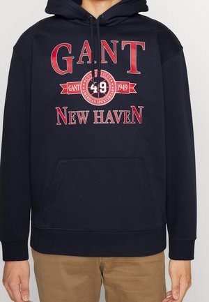 Sudadera de color azul marino hecha de una mezcla de algodón, con un bolsillo canguro en la parte delantera, capucha con cordón y texto gráfico rojo: "GANT 49 NEW HAVEN."