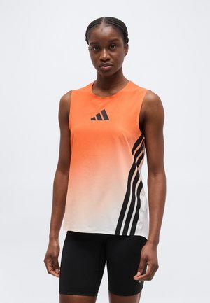 Žena nosi športový tielko s bezrukávovým dizajnom v oranžovo-bielom prechode so sivými pruhmi a čierne cyklistické šortky, stojaca pred jednofarebným pozadím.