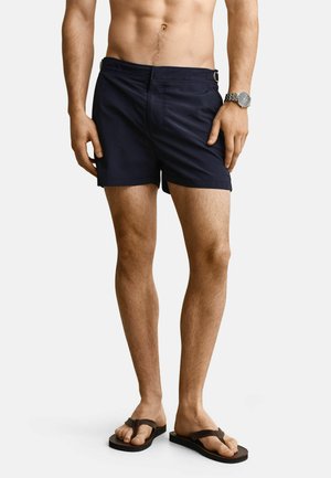 Torso et jambes d'un homme portant un short bleu marine, des tongs marron et une montre-bracelet argentée sur un fond uni.