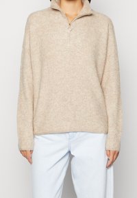 Beige gestrickter Pullover mit einem Halb-Reißverschlusskragen, lässiger Passform, gerippter Textur und langen Ärmeln, kombiniert mit hellblauen Jeans.