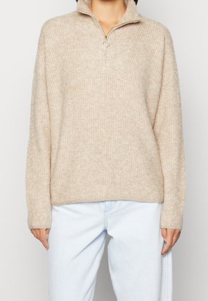 Beige gestrickter Pullover mit einem Halb-Reißverschlusskragen, lässiger Passform, gerippter Textur und langen Ärmeln, kombiniert mit hellblauen Jeans.