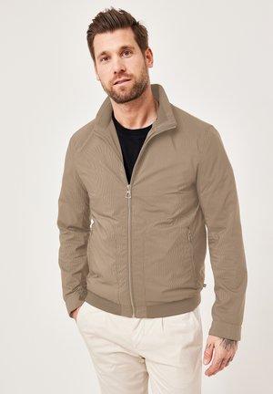 Light brown zip-up jacket with a ribbed texture, standing collar, side pockets, and fitted cuffs, worn over a black shirt.
