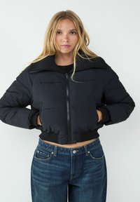 Czarna, krótka puchowa kurtka z zamkiem błyskawicznym i szerokim kołnierzem, z prążkowanymi mankietami i dołem, noszona na niebieskich dżinsach z wysokim stanem.