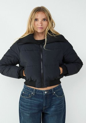 Zwarte cropped pufferjack met ritssluiting en brede kraag, met ribgebreide manchetten en zoom, gedragen over hoge blauwe spijkerbroeken.