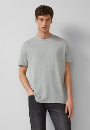 Młody mężczyzna o krótkich brązowych włosach, ubrany w jasnoszary krótkim rękawem t-shirt i ciemnoszare dżinsy, stoi na jednolitym szarym tle.