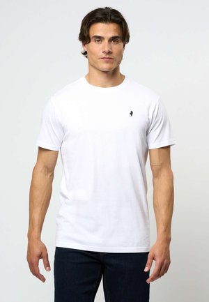 Bărbat purtând un tricou alb simplu cu mânecă scurtă și un mic logo negru pe pieptul stâng, asortat cu blugi bleumarin, stând în fața unui fundal simplu.