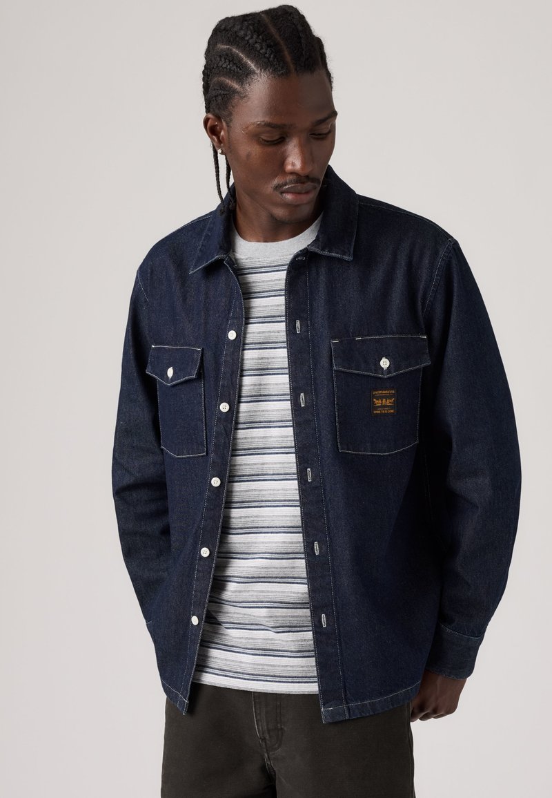 Jeune homme aux cheveux tressés portant une veste en jean foncée sur une chemise rayée grise et blanche, regardant vers le bas les mains dans les poches.