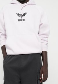 Person som bär en ljusrosa huvtröja med svart fjärilstrykt och japansk text, kombinerad med mörkgrå kordflorsbyxor med fickor.