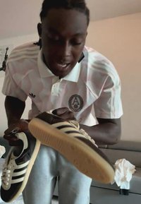 Person trägt ein weißes und pinkes Adidas-Shirt, hält ein Paar schwarz-weiße Adidas-Sneaker und zeigt sie drinnen.