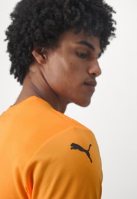 Orange atletiktrøje med struktureret finish og sort Puma-logo på skulderen. Designet har en moderne pasform og sporty stil.