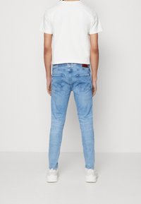 Vaqueros de mezclilla azul claro con un ajuste ceñido, bolsillos traseros y una pequeña etiqueta marrón en la cintura. Combinados con una camisa blanca de manga corta.