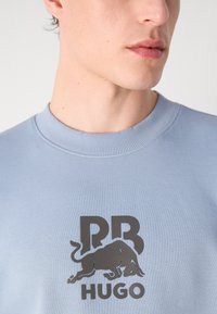 Sudadera azul claro con cuello acanalado, que presenta un gran logo negro de un toro y la palabra "Hugo" debajo. Textura suave.