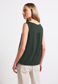 Ärmlös, mörkgrön top med rund halsringning, mjuk textur och samlingsdetalj på baksidan, i kombination med ljusa byxor.