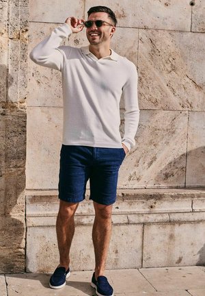 Man in wit shirt met lange mouwen, marineblauwe korte broek en instapschoenen, die zijn zonnebril rechtzet terwijl hij tegen een gestructureerde stenen muur staat.