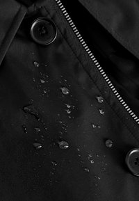Détail d'une veste noire imperméable comprenant un bouton, une fermeture éclair et des gouttes d'eau sur la surface texturée du tissu. Finition lisse et mate.