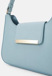 Sac à bandoulière en cuir bleu clair avec fermeture à rabat et plaque dorée gravée "ANNA FIELD" sur le devant.