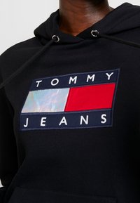 Sudadera negra con capucha que presenta un logotipo rectangular multicolor con secciones en azul, rojo y un blanco iridiscente, con texto bordado que dice "TOMMY JEANS."
