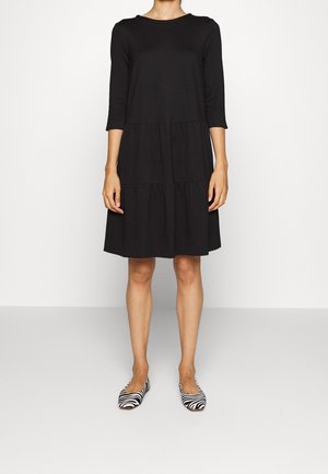 Femme portant une robe noire longueur genou avec des manches trois-quarts et des chaussures plates à motif zèbre, debout devant un fond blanc uni.