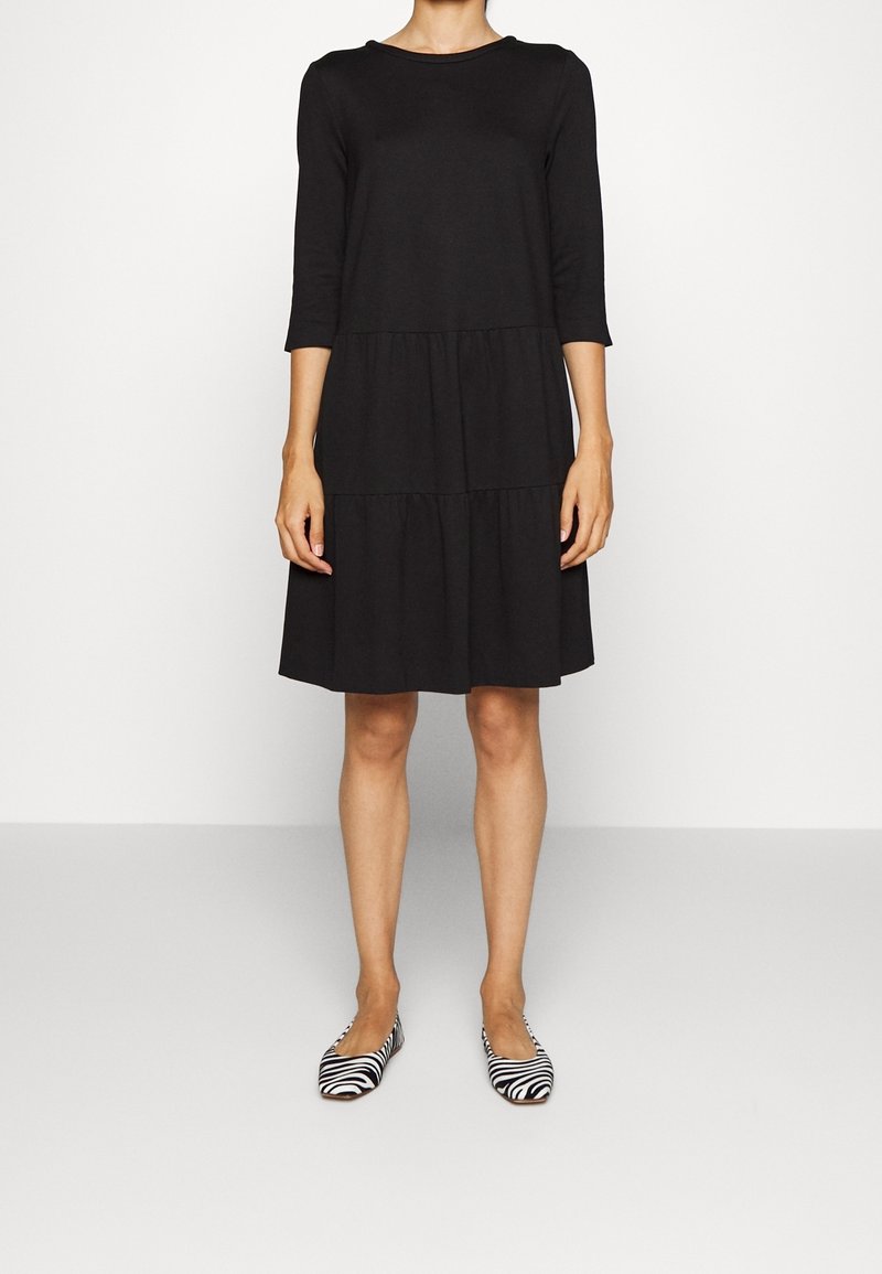 Femme portant une robe noire longueur genou avec des manches trois-quarts et des chaussures plates à motif zèbre, debout devant un fond blanc uni.