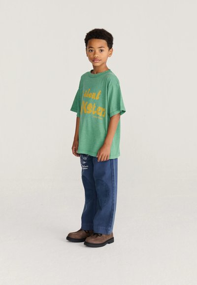 Jeune garçon debout portant un t-shirt graphique vert oversize, un pantalon bleu avec des patchs et des chaussures marron, sur un fond clair uni.