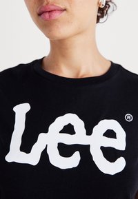 Melns kokvilnas T-krekls ar lielu, baltu, plūstoša stila "Lee" logotipu, apaļa apkakle un īsām piedurknēm.