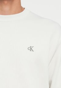 Hvit bomull sweatshirt med ribbet ribbkrage og liten grå brodert 'cK'-logo på venstre brystområde. Myk tekstur.