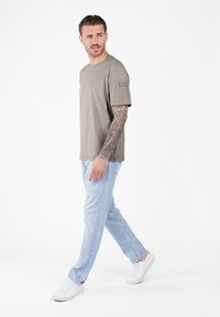 Hombre con un brazo tatuado caminando, vistiendo una camisa de manga corta color topo, jeans azul claro y zapatillas blancas, mirando hacia su izquierda.