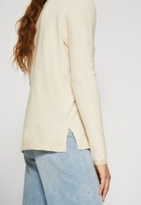 Femme aux longs cheveux bruns portant un pull crème à manches longues et un jean bleu clair, vue de dos à hauteur de la taille.