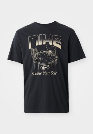 Melna kokvilnas t-krekls ar grafisku druku, kurā attēlots cilvēks karstā vannā, ar tekstu "NIKE" un "Soothe Your Sole" gai�ši zeltītā burtnīcā.