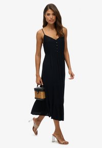 Zwart, mouwloos maxi-jurk met een gelaagd ontwerp, met knoopaccenten aan de voorkant. Doorzichtige haksandalen en een kleine rieten handtas.