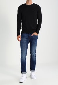 Schwarzer Pullover mit langen Ärmeln, taillierte Passform, Rundhalsausschnitt. Blaue Jeans mit leichtem Farbverlauf, hochgerollte Bündchen. Weiße Sneaker mit Akzenten.