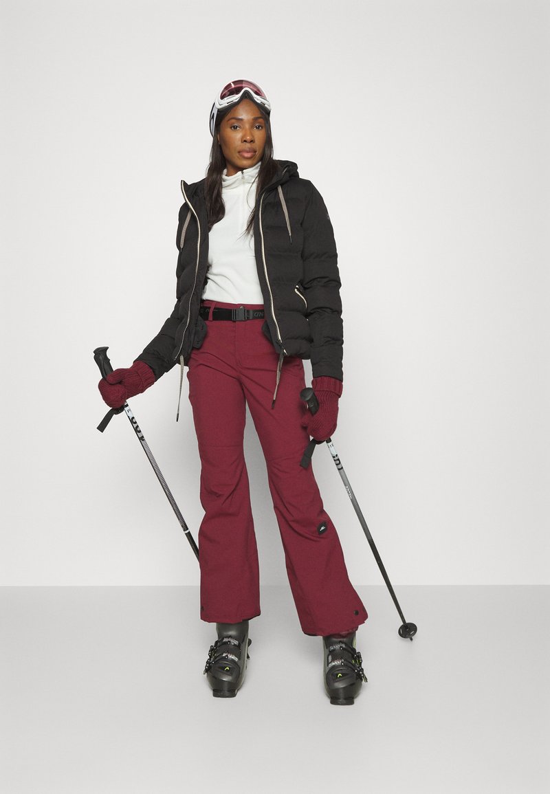 Czarna kurtka puchowa, biała bluzka, burgundowe spodnie narciarskie, czarne rękawice. W rękach kijki narciarskie, na głowie gogle. Czarne buty narciarskie z paskami.