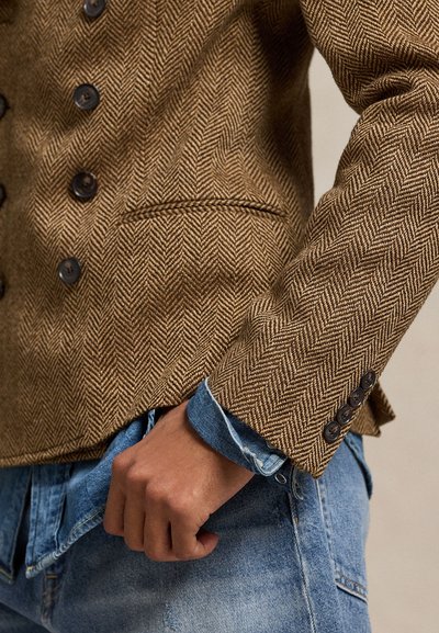 Καφέ σακάκι με σχέδιο ψαροκόκκαλο, μαύρα κουμπιά, μία τσέπη στο στήθος και λεπτομέρειες στα μανίκια. Φορεμένο πάνω από ένα ανοιχτό γαλάζιο πουκάμισο από τζιν.