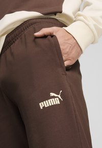 Braune Sweatpants mit elastischem Bund, seitlicher Tasche und einem hellen Puma-Logo auf dem Oberschenkel. Glatte Textur, lässiges Design.