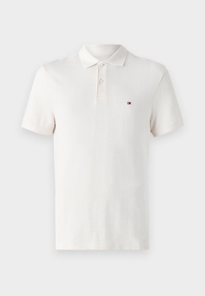 Polo majica v kremni barvi z teksturiranim črtastim vzorcem, z gumbom za zapenjanje na tri gumbe in majhnim logotipom na levem prsnem delu. Kratki rokavi.