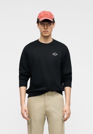 Ung mand iført en sort sweatshirt med et lille hvidt logo, beige cargobukser og en falmet rød baseballkasket, stående mod en ensfarvet baggrund.