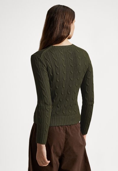 Alyvuogių žalios spalvos megztinis su pynimo raštu, aptempto silueto, su raštuota tekstūra ir ribomis apačioje bei rankogaliuose, dėvimas su rudomis kelnėmis, matomas iš nugaros.