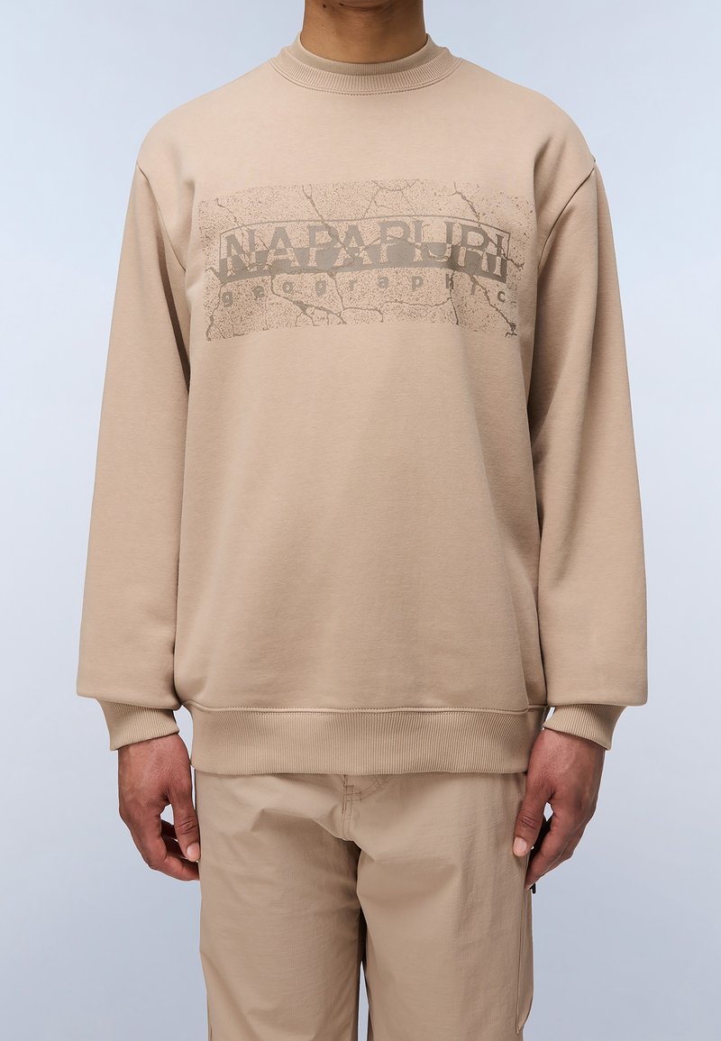 Homme portant un sweat-shirt et un pantalon beige, le sweat-shirt présente un texte "NAPAPIJRI" délavé avec un motif de texture craquelée sur la poitrine.