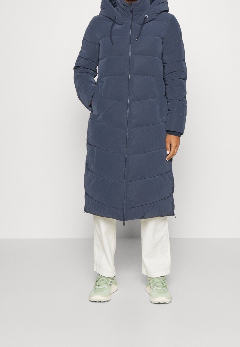 Lang navy puffer jakke med hætte, med lynlås lukning og horisontal quiltet design. Kombineret med hvide bukser og lysegrønne sneakers.