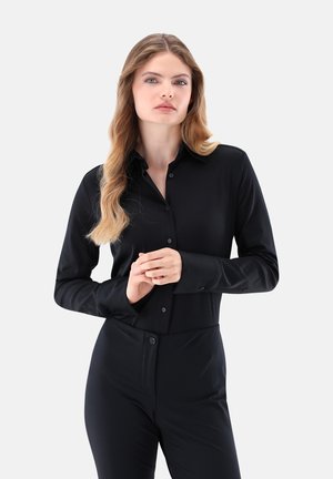 Schwarzes Langarmshirt aus glattem Stoff, mit Knopfleiste, spitzem Kragen und Ein-Knopf-Manschetten. Das Modell posiert mit gefalteten Händen.