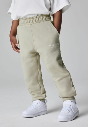 Kind dat lichtgroene sweatpants draagt met elastische tailleband en "ALLSAINTS" logo, witte sneakers en een wit T-shirt met korte mouwen.